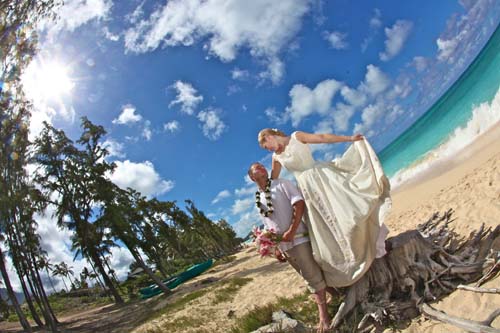 Bride lifting her gown beside driftwood on a sunny beach with the groom